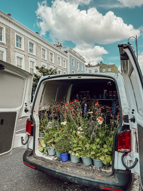The open rear of a delivery van filled with potted flowers and garden plants, including red and white blooms and green foliage, arranged closely together on the floor of the vehicle. The potted plants