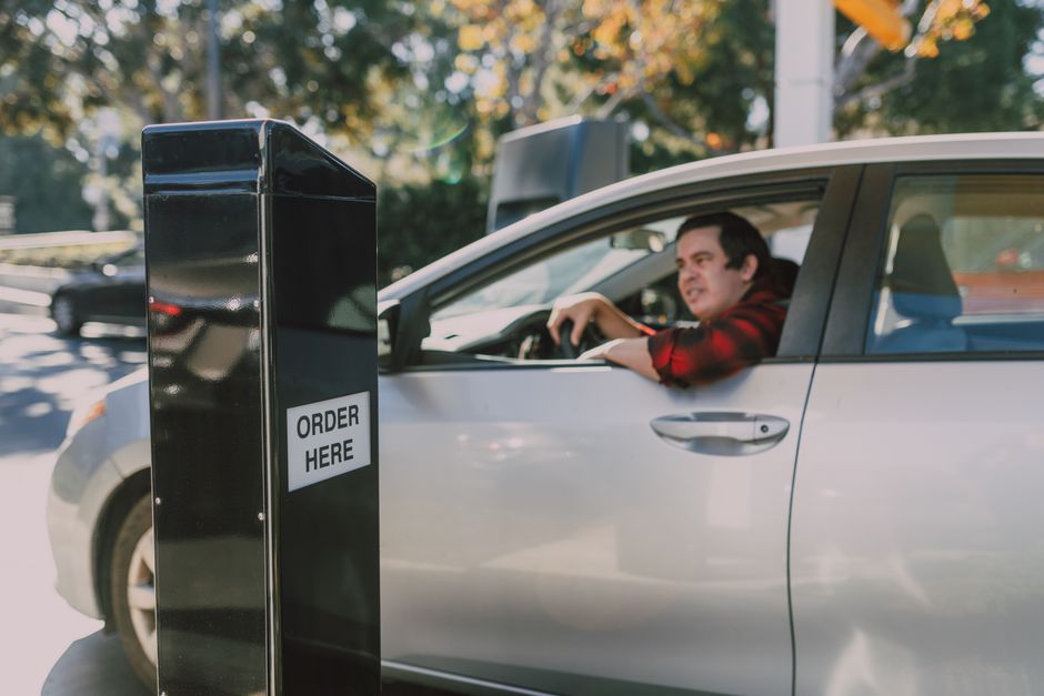 A young man experiencing mild frustration is seated in the driver’s seat of a silver car, positioned at a curbside flower order pick-up station next to a tall black stand with a white label reading 