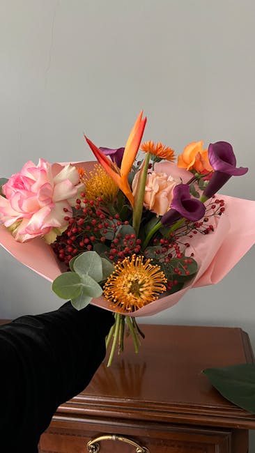 A vibrant floral bouquet featuring a mix of fresh flowers including pink carnations, orange gerbera daisies, purple calla lilies, and a striking orange bird of paradise. The arrangement is accented wi