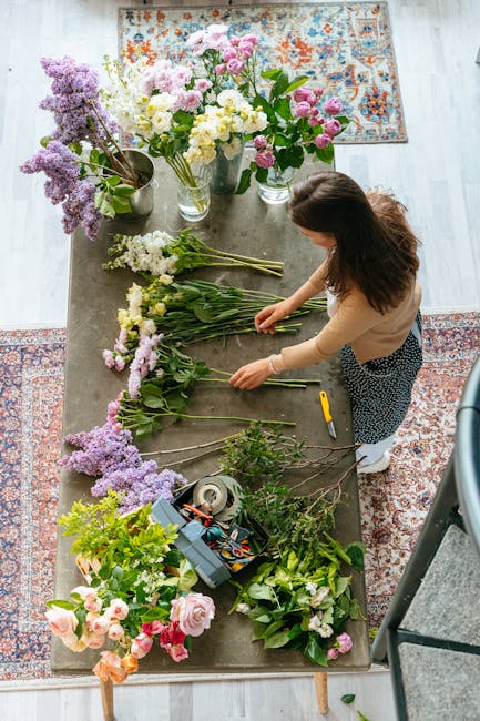 A woman arranging a variety of fresh flowers on a wooden table in a well-lit room, with bouquets including lilacs, roses, and other pastel-colored blossoms. The flowers feature shades of pink, white, 