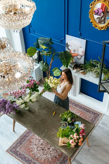 A woman arranging a floral bouquet inside a flower shop with vibrant blue walls, surrounded by various fresh flowers including white roses, pink roses, and purple blooms. She is holding a bouquet of w