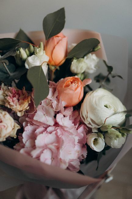 A close-up of a floral bouquet featuring soft pink hydrangeas, white ranunculus, peach roses, and light pink carnations, accented with dark green eucalyptus leaves. The bouquet is wrapped in light pin