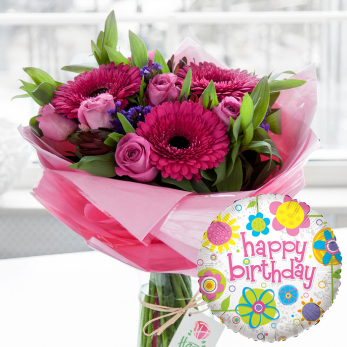 A sumptuous birthday bouquet with a matching foil balloon fills the frame, radiating joy and warmth the way a surprise delivery might brighten a home off Chislehurst High Street. The arrangement sits proudly in a clear glass cylindrical vase, its stems visible through the water, emphasising the freshness of each flower. At the centre, large gerbera daisies in rich tones of fuchsia and magenta create a striking focal point, their petals perfectly layered around dramatically dark central disks. These confident blooms are surrounded by clusters of soft pink and blush roses, some still tucked into tight buds and others gently opening into cupped blossoms, their spiralling petals suggesting tenderness and affection. Sprigs of deep purple filler flowers, likely statice, thread through the design, adding depth and a slightly wild note that keeps the bouquet from feeling too formal. The whole arrangement is embraced by generous green foliage, with leaves of varying shapes and textures that frame the flowers and enhance the sense of fullness and movement. At the base, translucent light pink cellophane or tissue is wrapped around the stems, its softly crinkled surface catching the light and creating a delicate halo that echoes the pink tones of the blooms. A length of natural twine is tied in a simple bow around the neck of the vase, holding a small off-white gift tag decorated with a discreet rose-like emblem, quietly suggesting a premium touch from a skilled local florist. In the lower right foreground, partially overlapping the bouquet, a round Mylar Happy Birthday balloon adds a playful accent. The balloon has a white background flecked with grey, overlaid with colourful flower and dot patterns in bright yellow, sky blue, lime green, vivid pink, and deep purple. Bold pink cursive text spelling happy birthday arcs across its centre. Soft natural light, reminiscent of a bright morning in a Chislehurst dining room, bathes the scene and gently blurs the background, making this floral and balloon gift set the clear, joyful focus of the image.